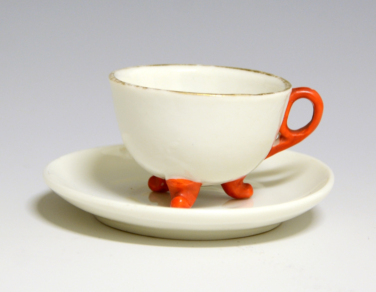 Kopp og skål til dukkeservise i fajanse, hvit glasur med gulldekorerte kantrender. Koppen har rød hank og står på tre føtter som også er malt røde.