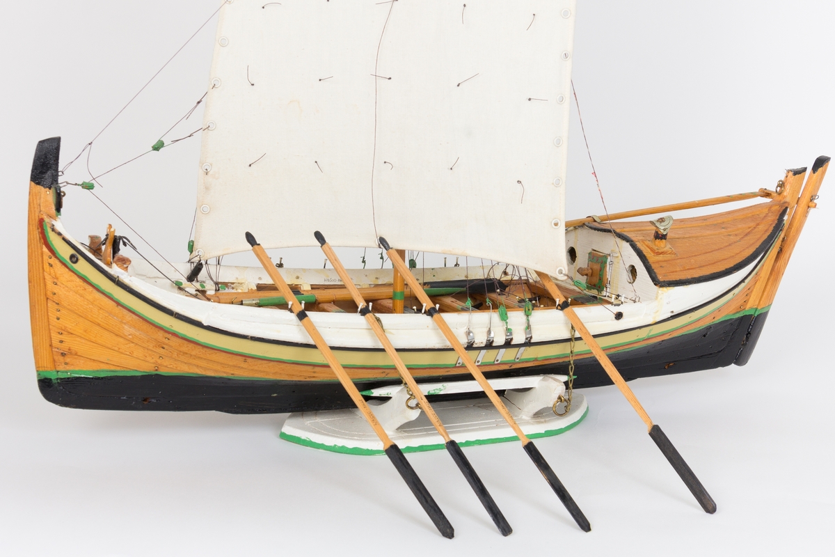 Modell av nordlandsbåt - en fembøring med syv årepar og fem par tollpinner, seks tofter og et øsekar og en båtshake. Båten har en løfting med pipe og dør og to ventiler i simsen og er rigget med råseil og toppseil. Ripa er malt rød, hvit, grønn, olivengrønn og sort. Kjølen og nederste delen av skroget er sortmalt (bunnstoff) med en smal grønn stripe øverst. Modellen står i en krybbe. I framskottet ligger det en dregg med fem klør. I tillegg har modellbyggeren limt fast 10 ulike skjell i framskottet. Ifølge informant ble denne typen dekor et trekk som kjennetegnet modellbyggerens båter etter hvert som han ble eldre.