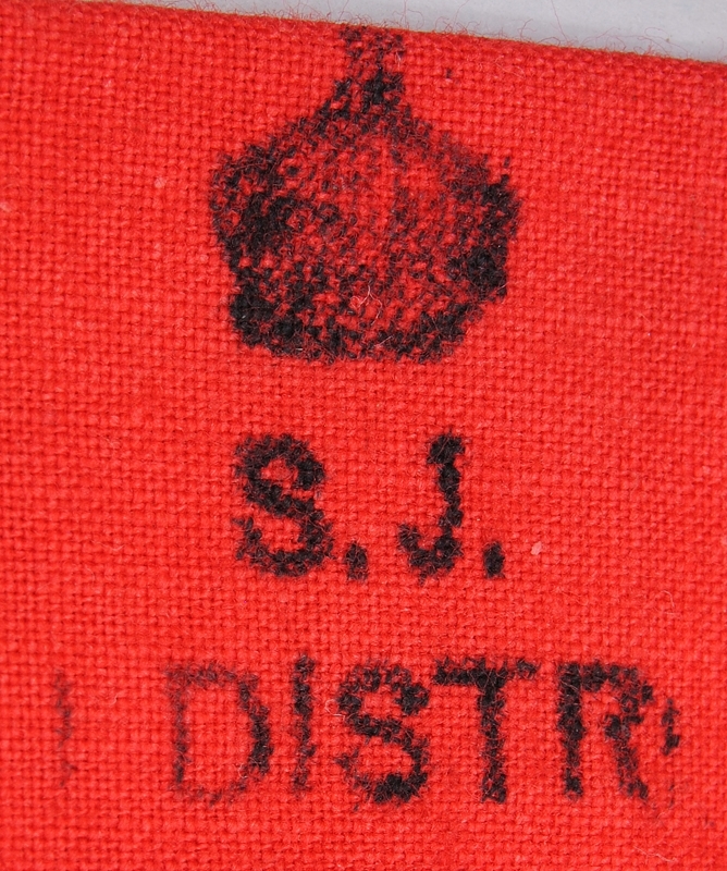 Röd signalflagga utan skaft. I kanalen som är sydd för skaftet finns texten: "S.J. 1 DISTR" tryckt och ovanför syns en tryckt illustration av en krona.