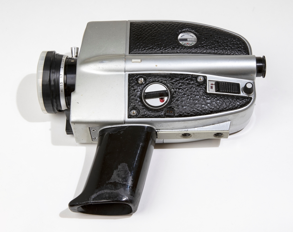 Filmkamera, super-8 BAUER C ROYAL 6-zoom med kamerahus av grå metall delvist klädd med svart skinnimitation. Handtaget är av svart plast och har utrymme för batterier och tillsluts av en lös platta. På linsen sitter ett genomskinligt linsskydd. Filmkameran är placerad i en svart väska, JM 56586, i vilken finns en fjärrutlösare, ljusskydd av gummi för linsen, liten gummiring för sökaren, och större gummiring.