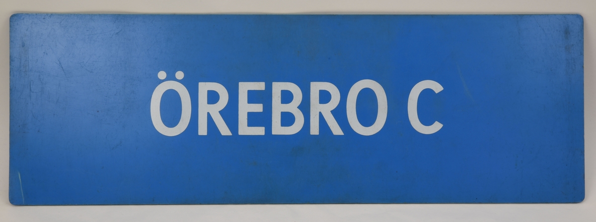 En blå avlång dubbelsidig destinationsskylt som har den vita texten "ÖREBRO C" på båda sidor.