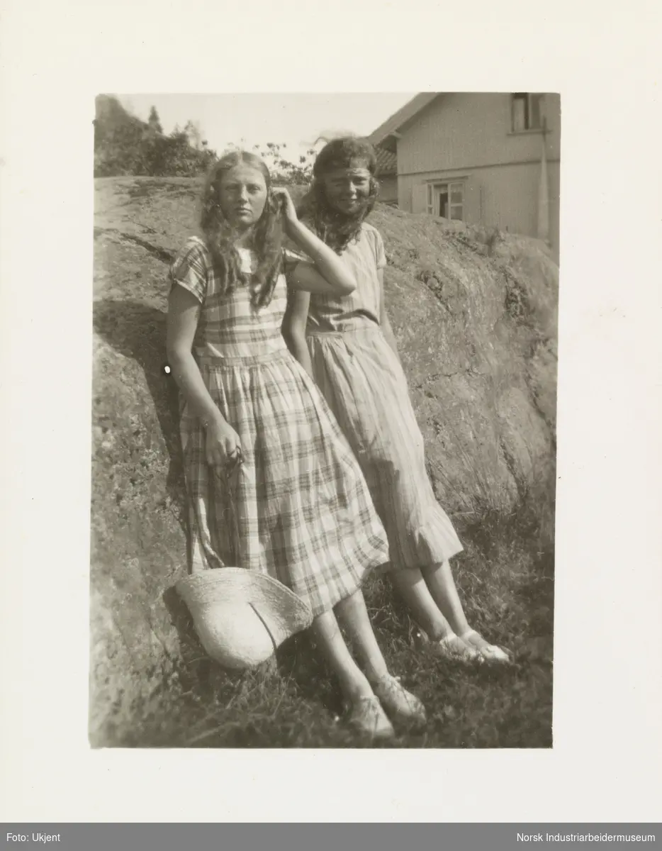 To jenter i sommerkjoler lener seg mot fjellside. Jentene har løst krøllete hår og sommersko