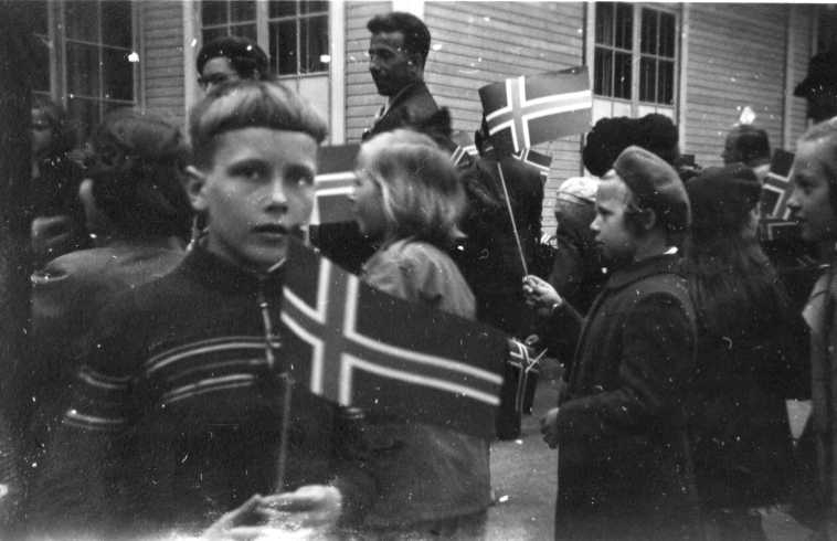 En folksamling med bland annat flickor och pojkar som viftar med norska flaggor. En pojk med flagga i handen är vänd mot fotografen.