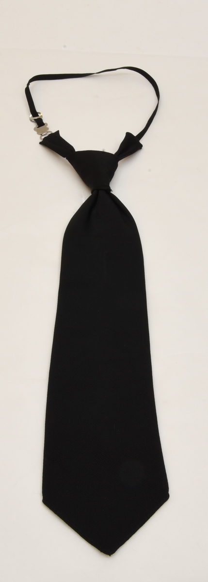 Färdigknuten slips av svart textil. Vid slipsens knut sitter två flikar av textil med spänne och resårband fäst i vardera del. Resårbandet träs runt nacken och fästs med hjälp av ett metallspänne.