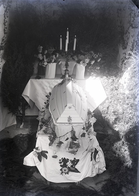 En ljus likkista står på en avsats täckt med ett ljust lakan, framför ett bord med duk, fem ljus och blommor.