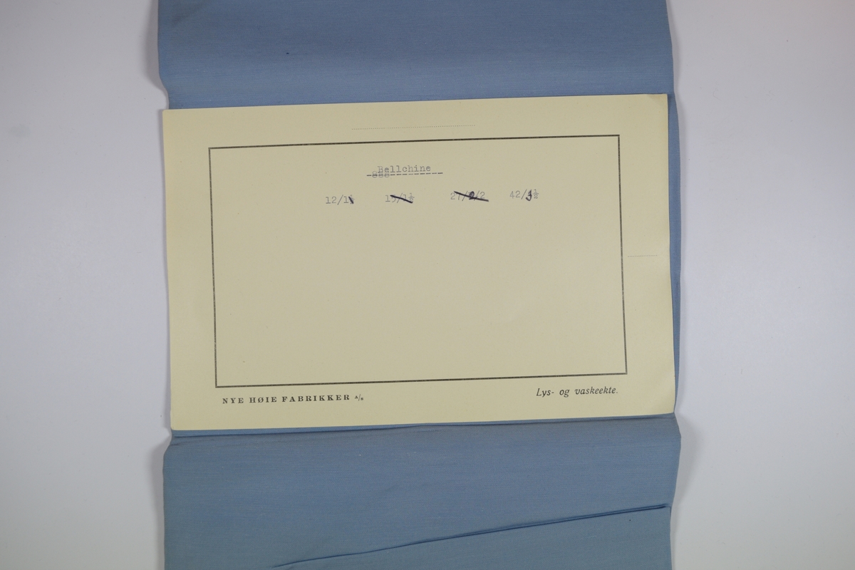 Tekstilprøve av sammenfoldet stoff, med påklistret papirlapp. Ensfarget tynt blått tekstil. Et papirkort ligger inni tekstilet.