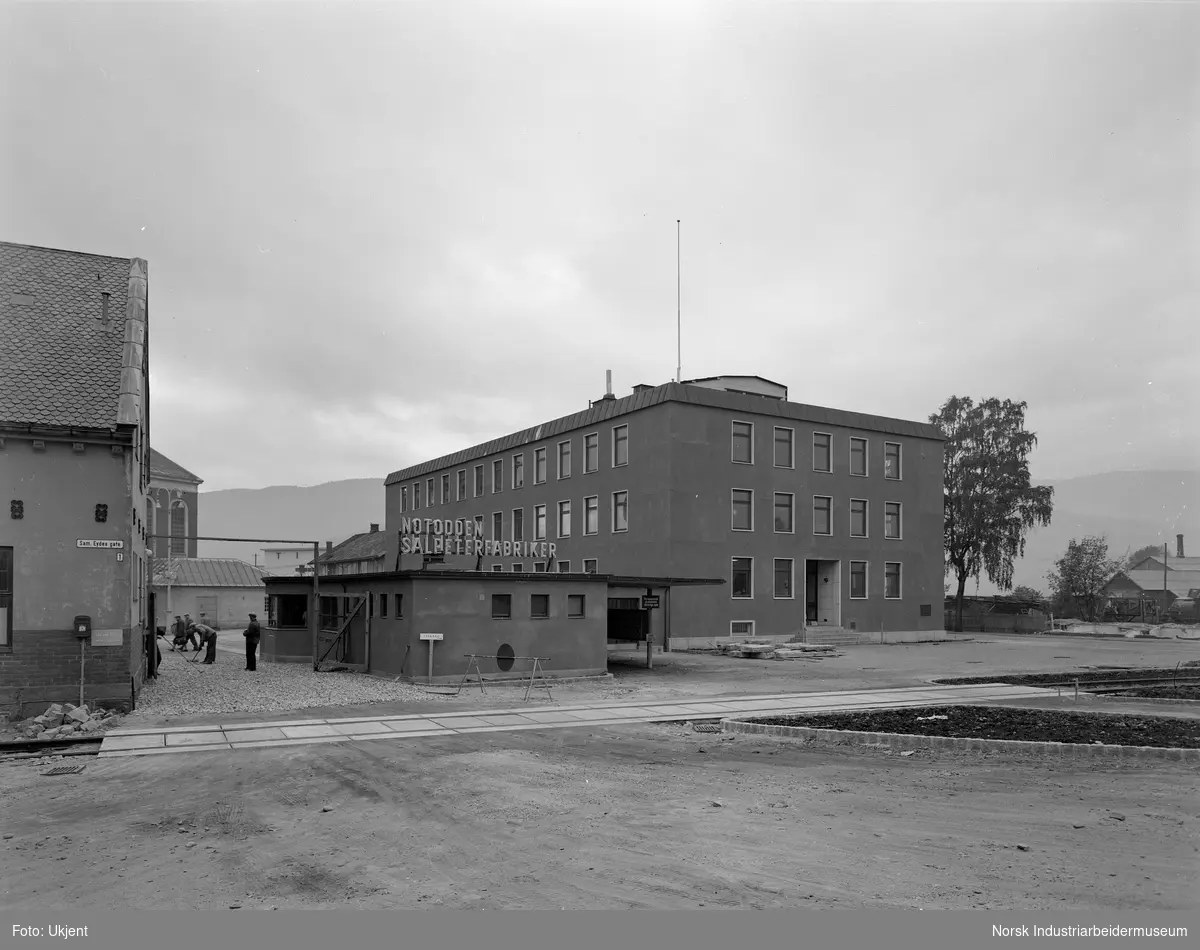 Arbeid utenfor portvakta som er arbeidernes inngang til fabrikken. Kontorbygget i Hydroparken til høyre (bygg 1, oppført 1955).