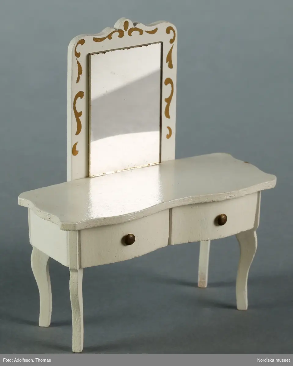 Toalettbord med spegel och gyllene dekor. Vitmålad trä, troligen tillverkad av Lerro. Hör till inredningen i föräldrarnas sängkammare på andra våningen i dockskåpet.