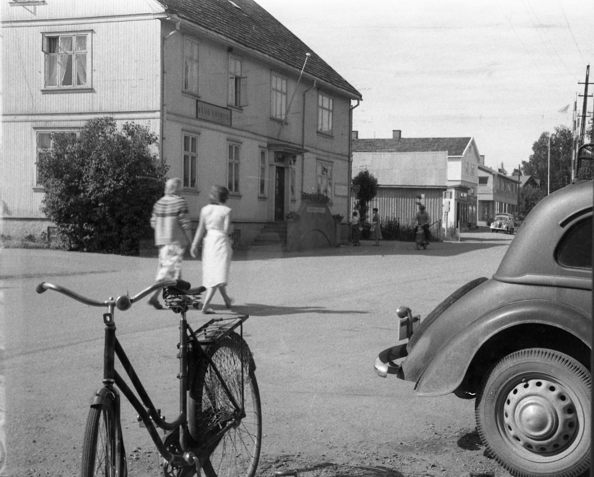 To gatebilder fra Lena sommeren 1958. Bildene er tatt gjennom vinduet fra Lena Foto & Radio i "Raubua" i retning Grand og jernbaneovergangen. Noen uidentifiserte personer går i gata. I høyre bildekant skimtes bakparten på en bil som iflg informant er en Adler Trumph Junior årsmodell 1934-40.  I forkant står en sykkel.