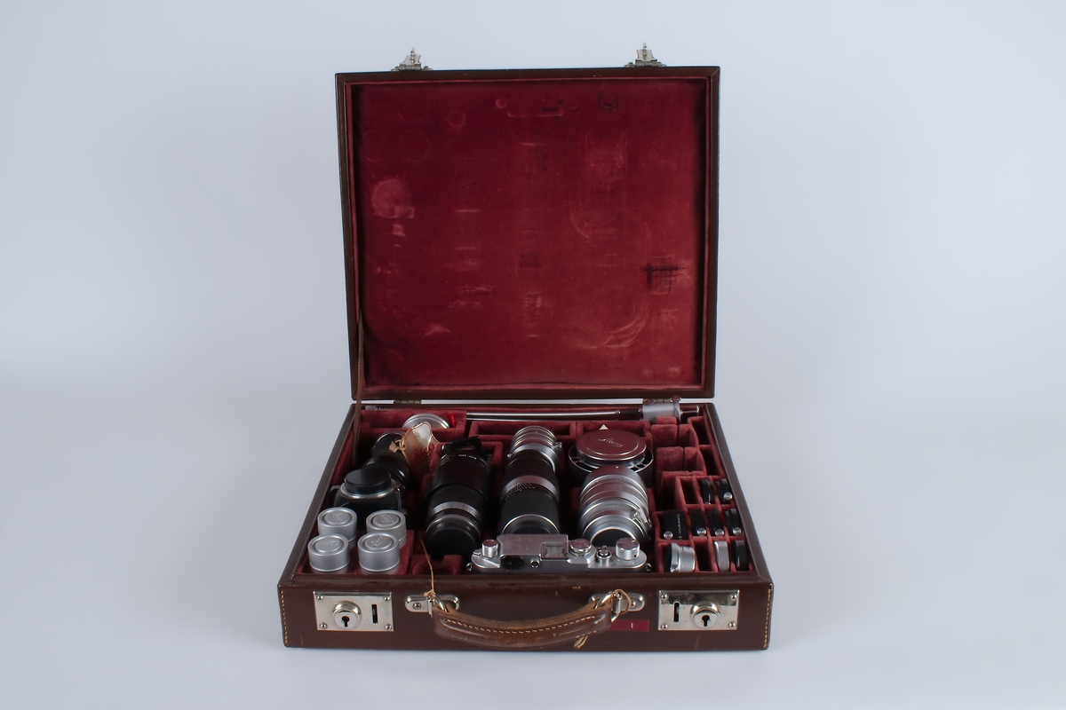 Fotografiapparat i brun koffert med rødt fløyelsfor. Kofferten inneholder i tillegg til selve kameraet en mengde ekstrautstyr.