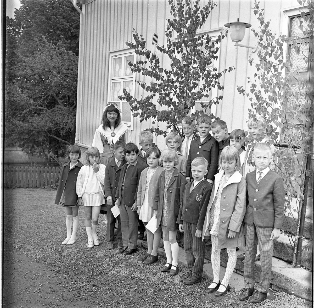 Skolavslutning med små barn i Ölmstad, sista skolklassen? Lärarinnan som bär folkdräkt heter möjligen Frilund. De står finklädda nedanför en trappa pyntad med björkris.