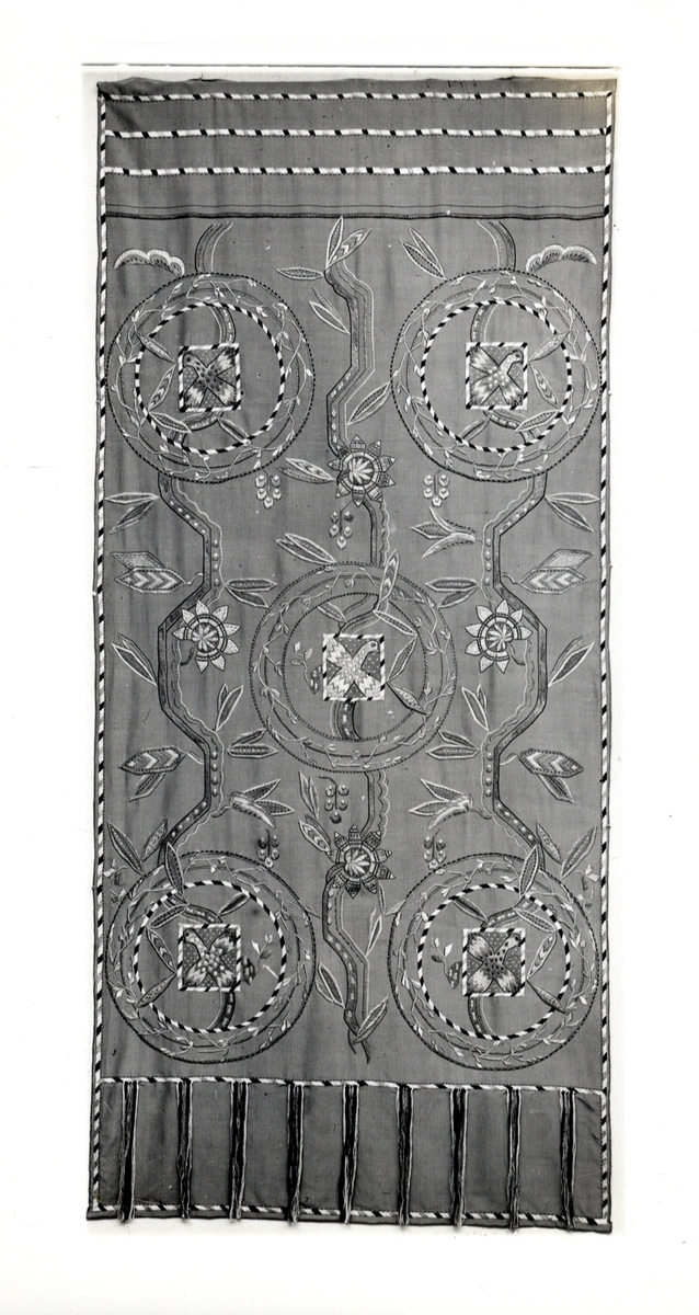 Foto (svart/vitt) av ett draperi broderat på siden, med motiv av fåglar och stiliserade blommor. 

Inskrivet i huvudbok 1983.
