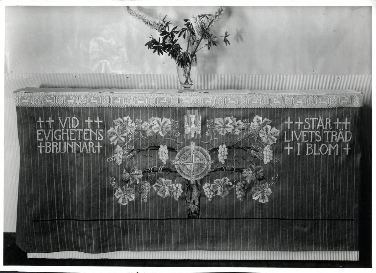 Foto (svart/vitt) av ett antependium broderat med Livets träd och text: "Vid Evighetens Brunnar Står Livets Träd I Blom".

Inskrivet i huvudbok 1983.
