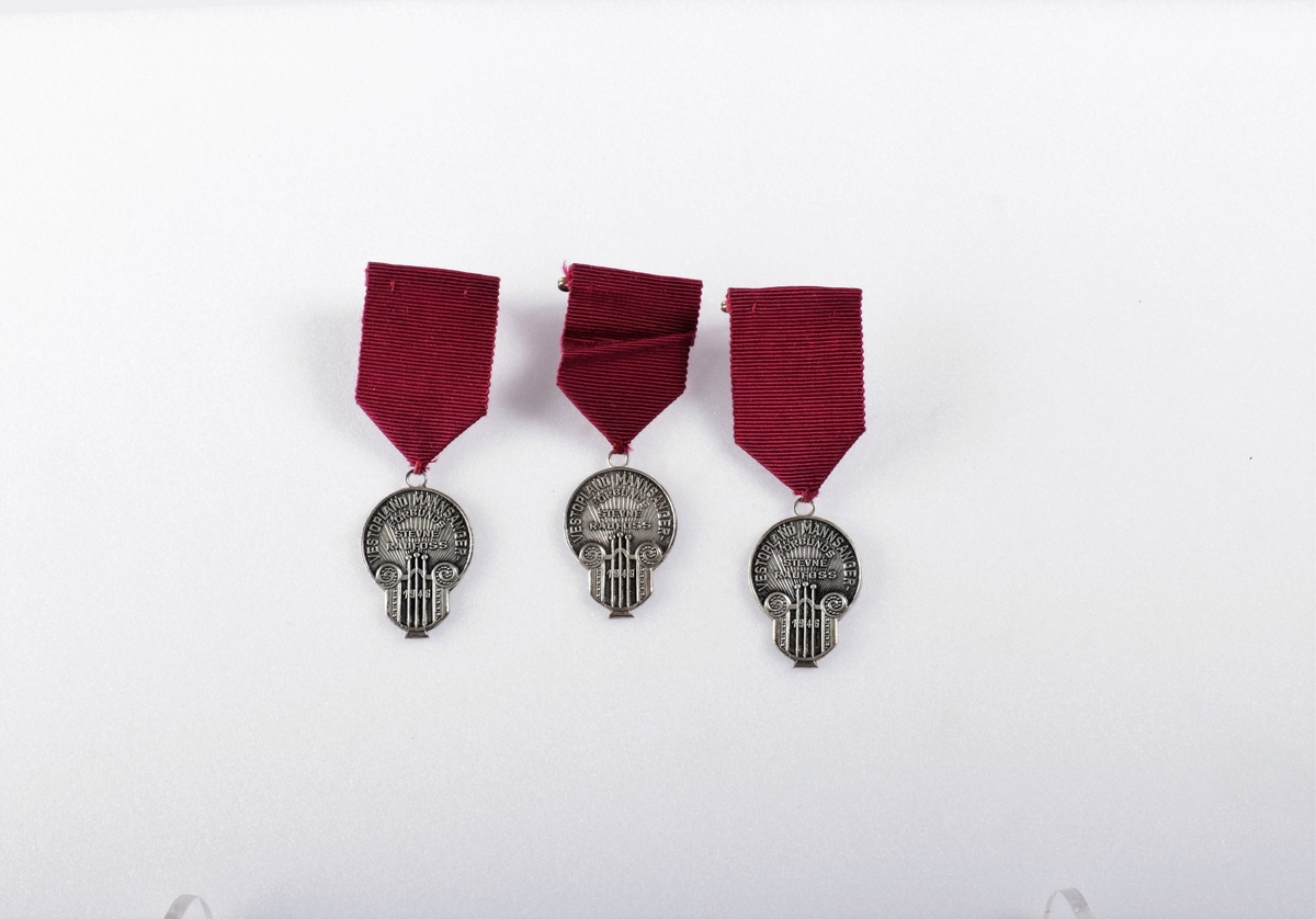 Tre like minnemedaljer av sølvfarget metall med purpur festebånd. Innskift på medaljen