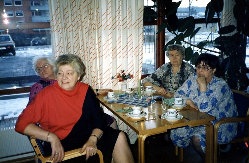 Fyra kvinnor fikar vid ett bord i Brattåsgårdens cafédel år 1989.
Från vänster: ? Johansson, (i rödsvart klänning) Sonja Hagman (1920 - 1999), okänd kvinna samt Inger Seger (1928 - 2009), Bölet. I bakgrunden till vänster skymtas Våmmedals bostadsområde.