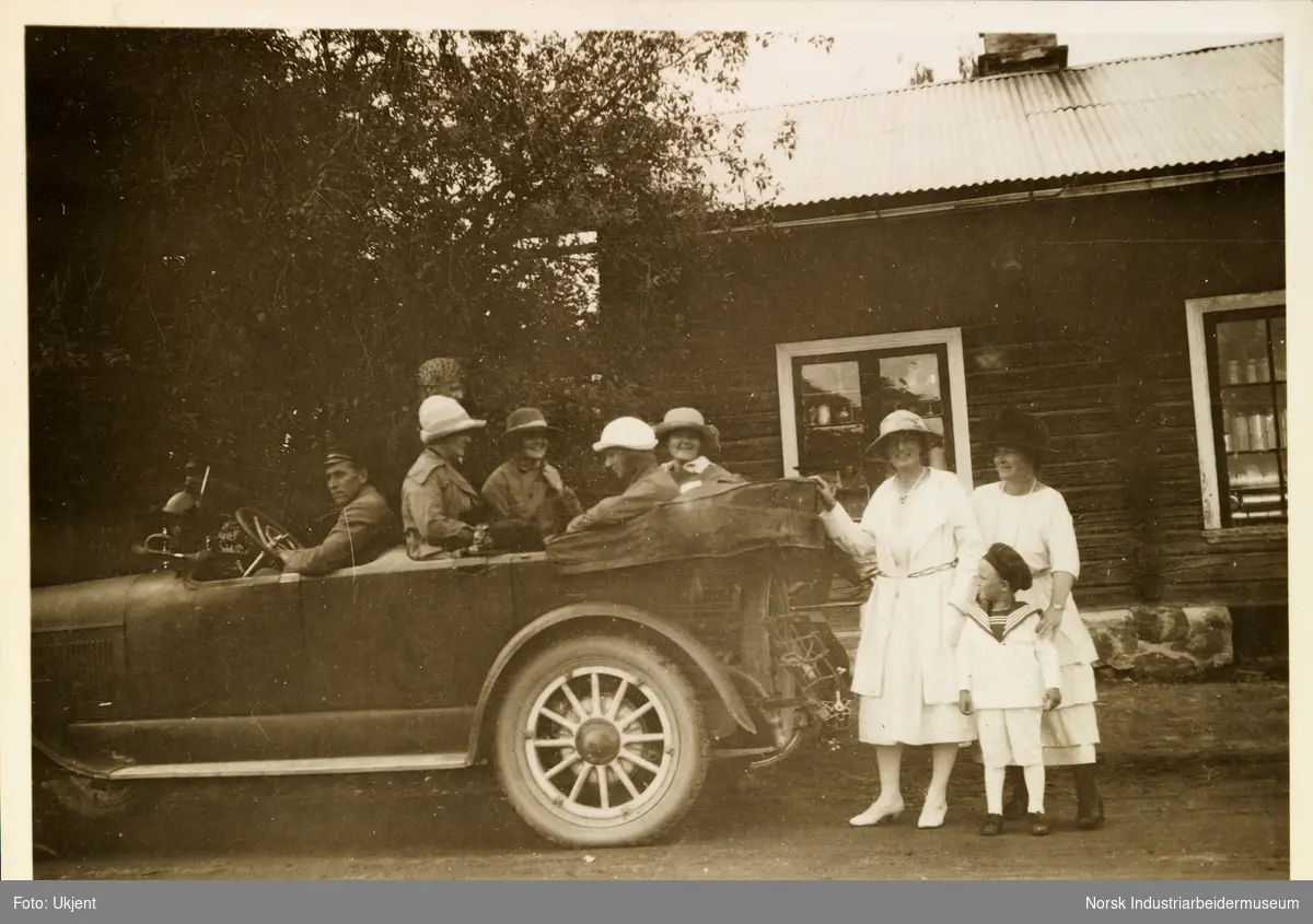 Mennesker sitter i åpen bil. Bak bilen står to kvinner og et guttebarn i lyse sommerklær. Bilen er plassert foran bolighus