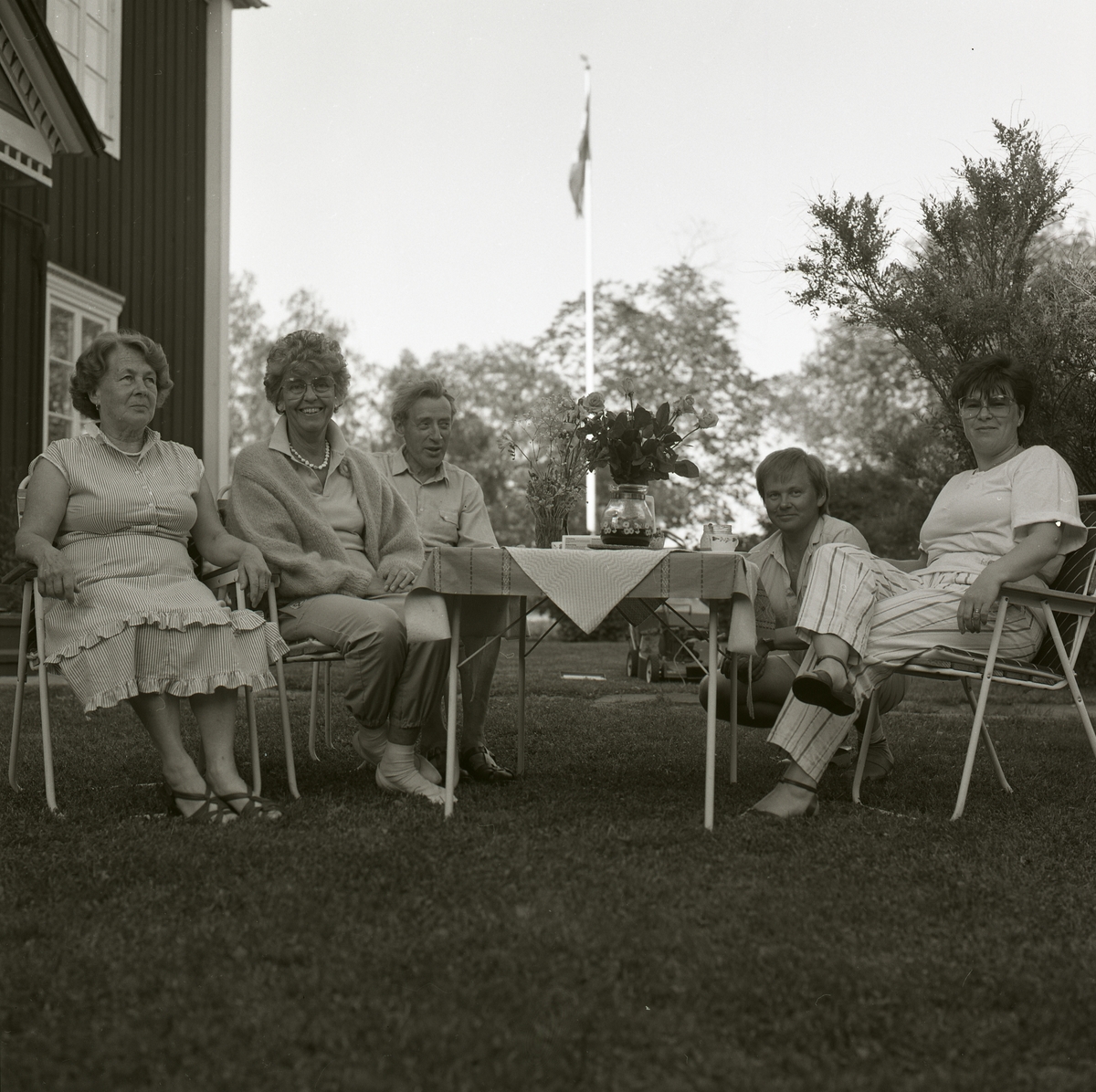 Adéle och några andra kvinnor och män sitter vid dukat kaffebord. På bordet ligger en duk och det står ett par blombuketter där. På sidan om står ett hus med snickarglädje och fönster, bakom är flaggan hissad. Det finns även träd och gräs. Adéles 68 årsdag juni 1986.