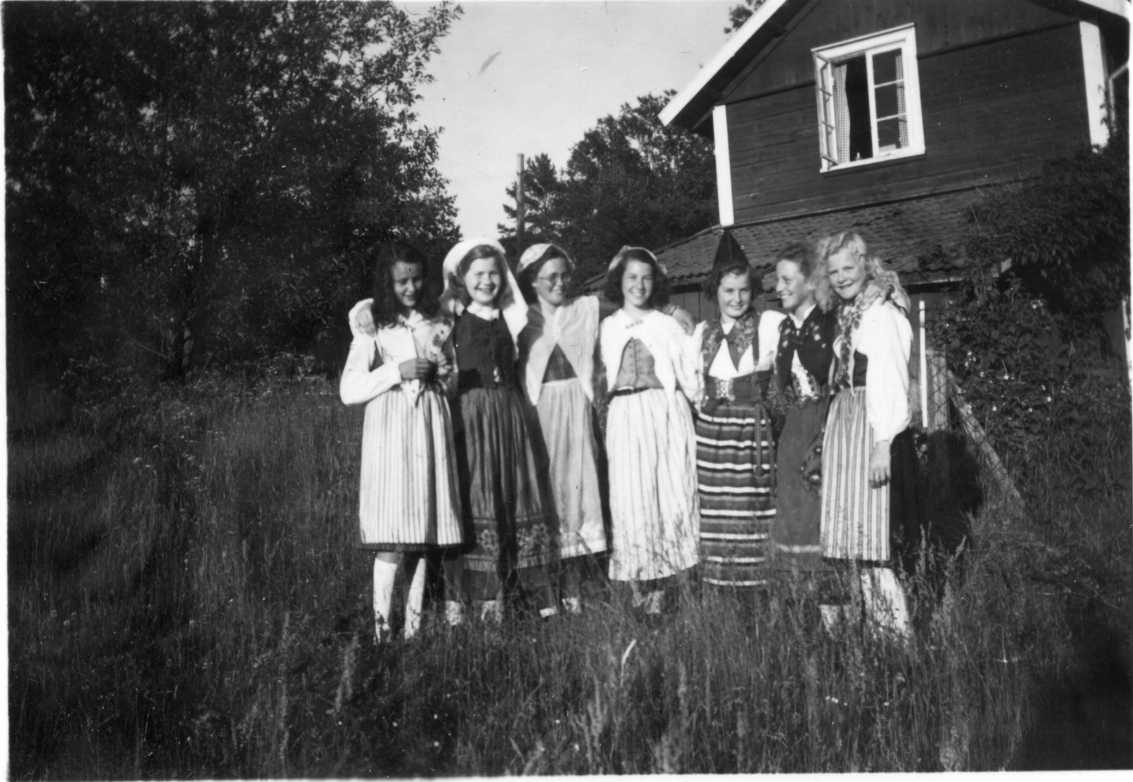 Sju flickor i folkdräkt står i högt gräs vid gavel till hus. Gaveln har en lägre tillbyggnad med pulpettak. Bland flickorna finns Ingrid Levén, Ulla-Britt Havström och Birgit Roman.