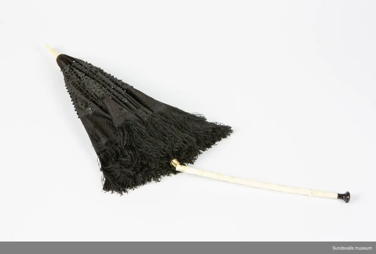 Svart parasoll med vikbart skaft i två material, trä och ev ben med skuren dekor. Svart knopp nedtill. Parasollet är broderat med svarta pärlor och försett med fransar.