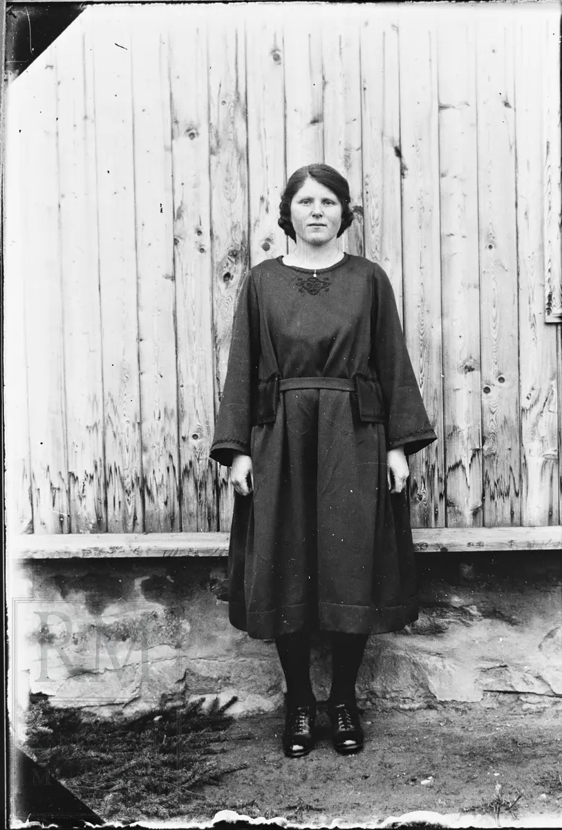 Portrett av ung kvinne, stående foran en husvegg, i mørk kjole.