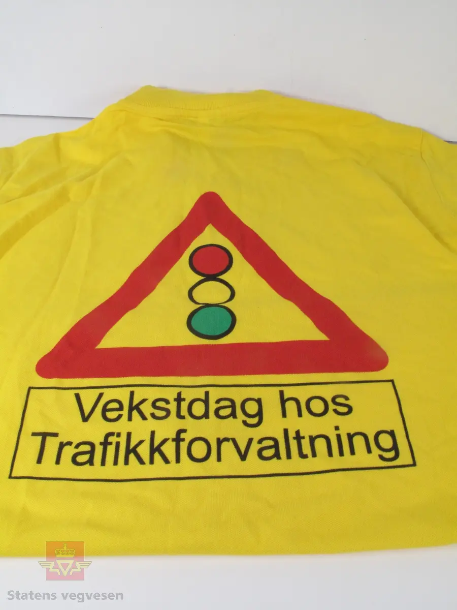 Gul t-skjorte (pique) med krage og knappelukking (3 knapper). Figur av et fareskilt med trafikksignal på baksiden, samt tekst. T-skjorten er størrelse Medium og tilnærmet ubrukt.