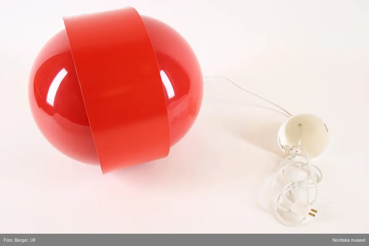 Taklampa, elektrisk, av röd blank akrylplast, stående oval form med ett förkroppat band av vinyl runt mitten. Vit plastsladd, konisk takkopp av vit plast.
/Maria Maxén 2017-06-27