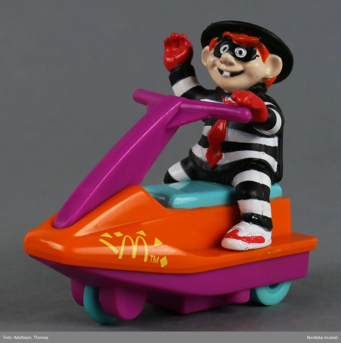Mekanisk leksak som föreställer en maskerad pojke i randig fångdräkt, slängkappa och vidbrättad hatt. Figuren sitter på en orange skoter med tre små hjul på undersidan, som går att dra upp så att fordonet rullar.