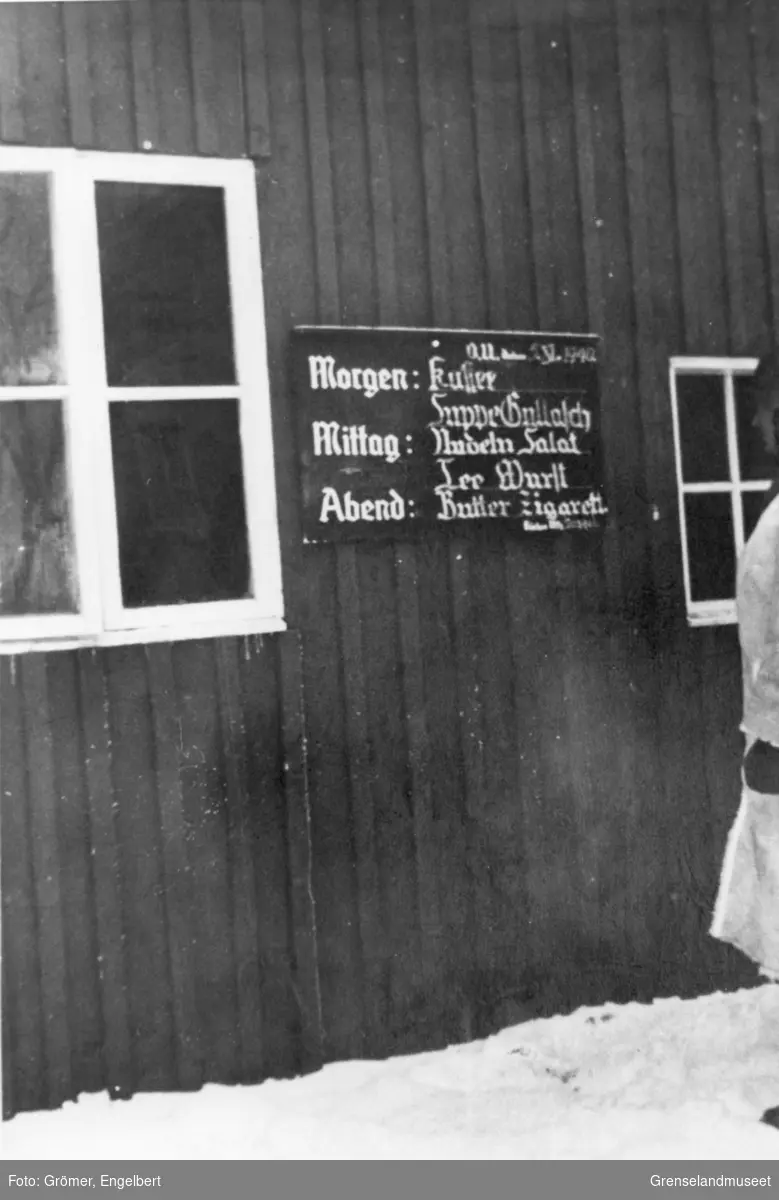 Et tyskspråklig skilt henger på utsiden av en bygning. Skiltet er merket: O.U. den 2 V. 1940 og det som ser ut til å være en oversikt over dagens måltider. En soldat i vinteruniform står og leser på skiltet.
