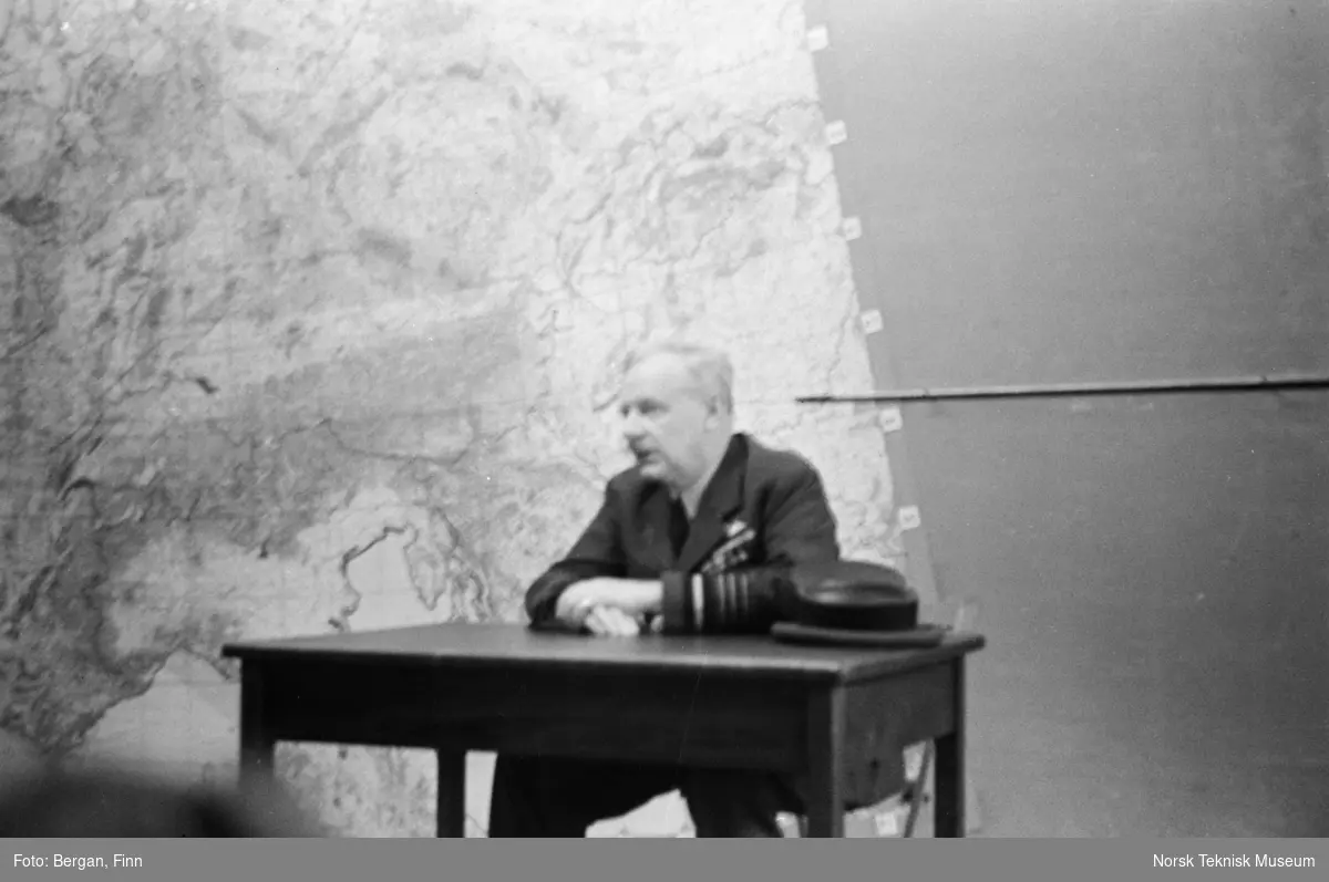 Person i uniform sitter ved bord foran stort kart uskarpt bilde