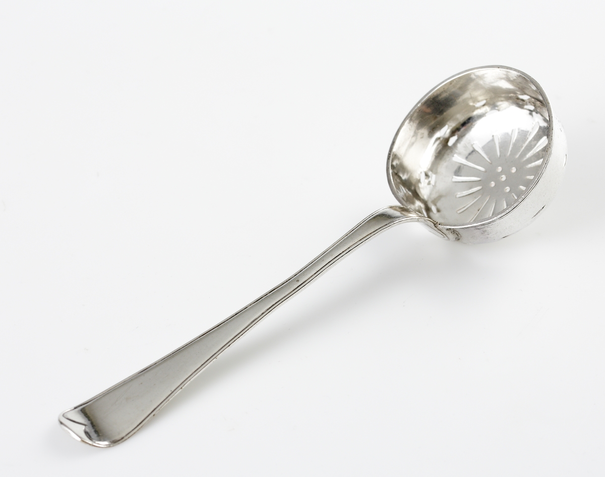 Strösked i silver.
Svensk trubbig modell. Perforerat runt skedblad med genombrutet strålformat mönster och fem runda hål. Längst ut två rader med avlånga genombrytningar. Ägarinitialer: "I.O.S." på baksidan av skaftet. Stämplar på baksidan av skaftet.