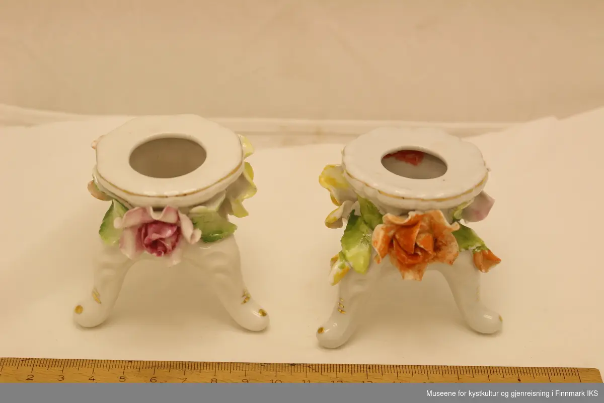 To små lysestaker i porselen. Lysestakene står på tre bein, og har dekorative blomster og blader rundt kanten, og med gullfarve malt rundt kanten og på hvert av bena.
