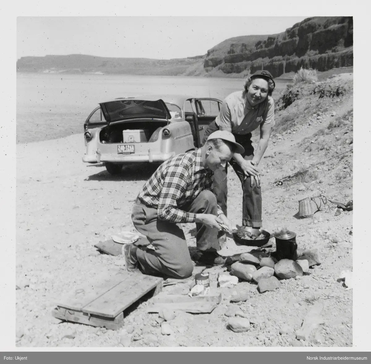 Mann og kvinne lager mat over bål på strand. I bakgrunnen er bil med åpne dører parkert på stranden