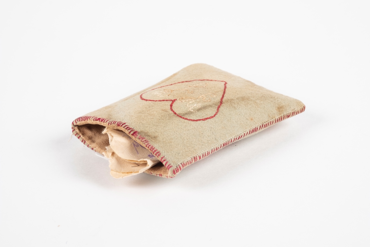 En liten firkantet lomme laget av papp kledd med tekstil. Inne i lommen er det en papirlapp. Det er brodert ruter med årstall under på den ene siden og et hjerte med bokstaver i på den andre.