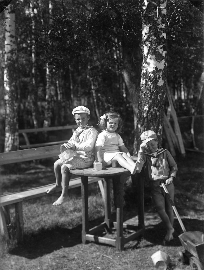 Barnen Stolpe från Stockholm. En liten flicka i ljus klänning sitter på ett trädgårdsbord intill en något äldre pojke. En annan pojke står lutad mot bordet och håller en leksakskärra i handen. Pojkarna bär sjömansuniform.