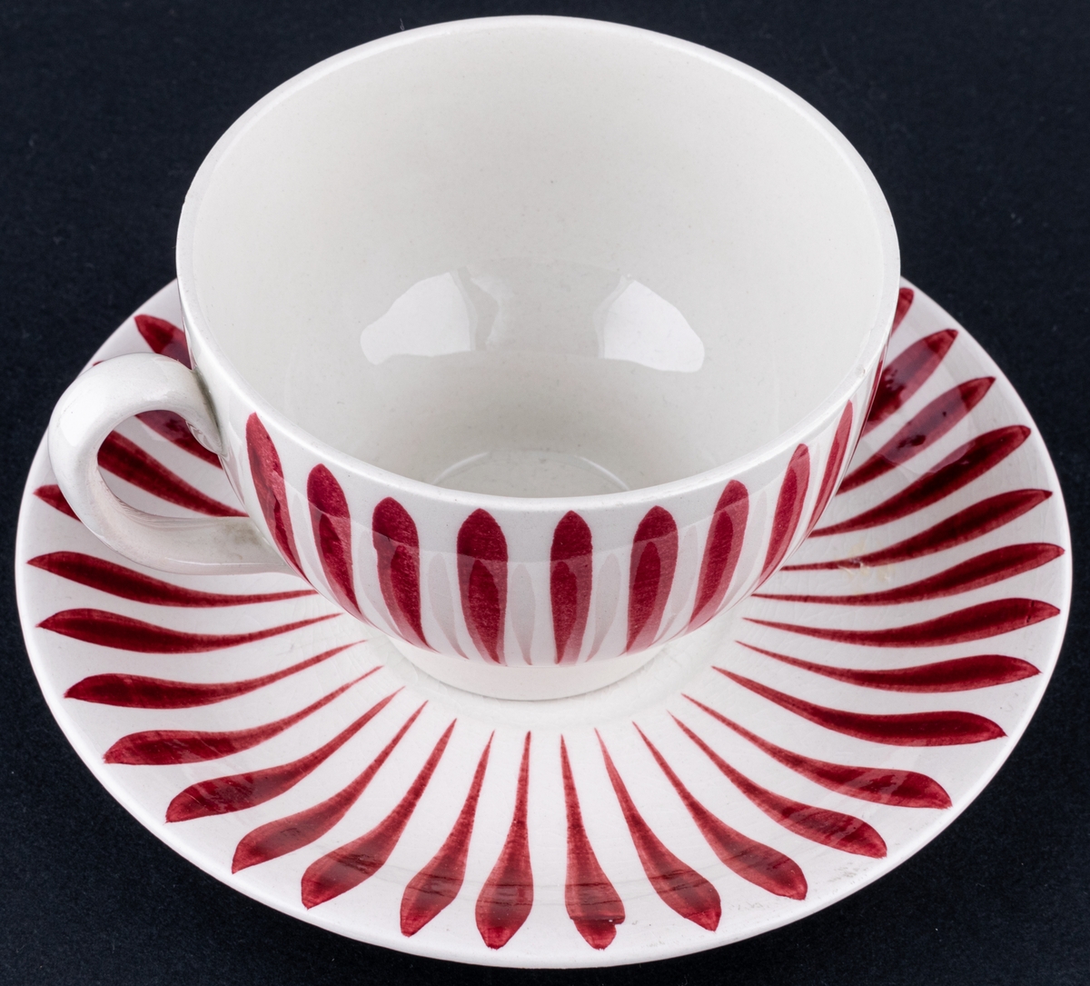 Kaffekopp med fat, modell AX i dekoren Aster. Finns i röd och blå dekor. Dekoren skapad av Eugen Trost. Dekoren runt koppen är i röda vertikala streck och på fatet dito runt brättet.