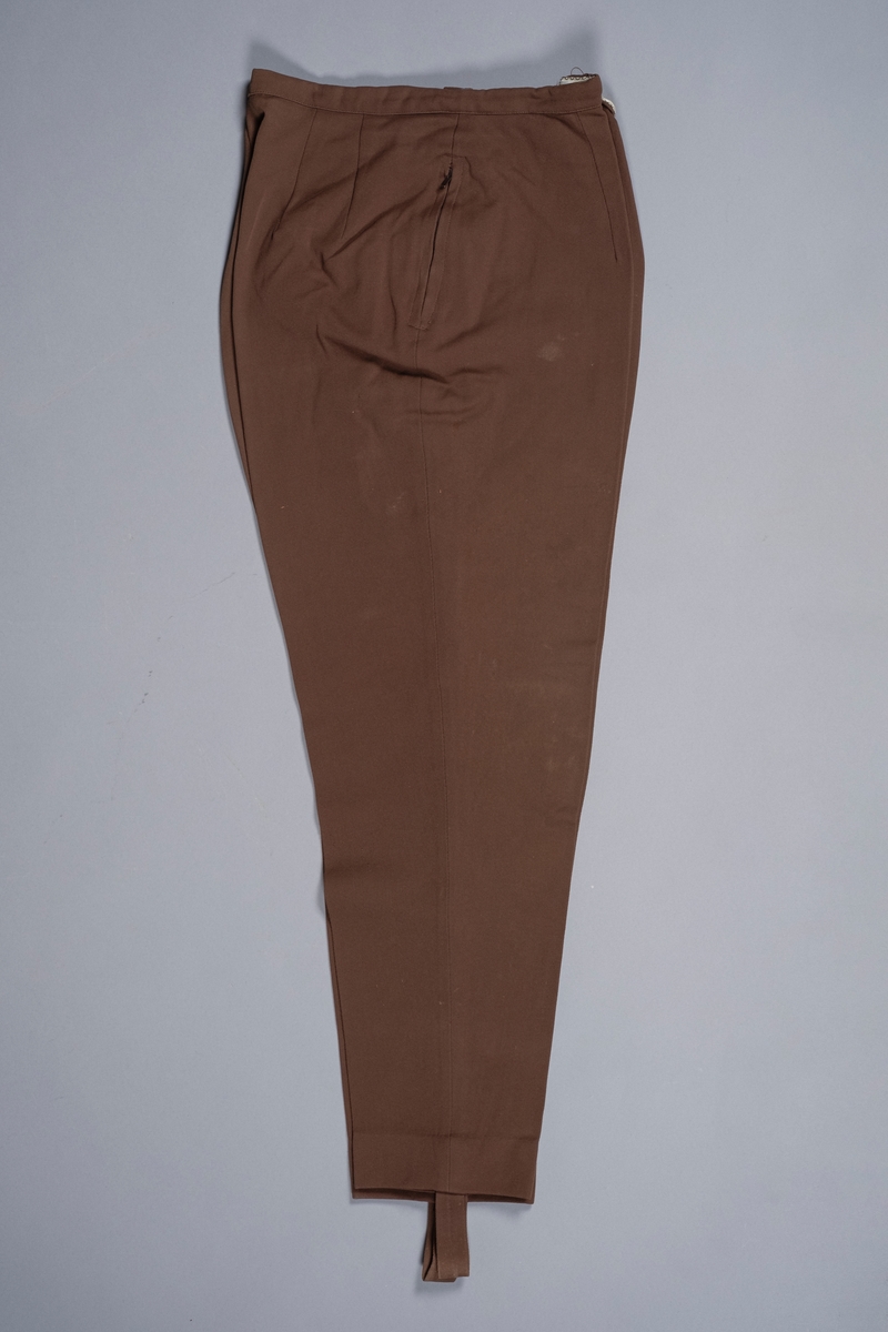 Bukse i brunt stoff som smalner nedover og ender med fotløkker. Buksen lukkes med glidelås i siden. På den andre siden er det en glidelåslomme.