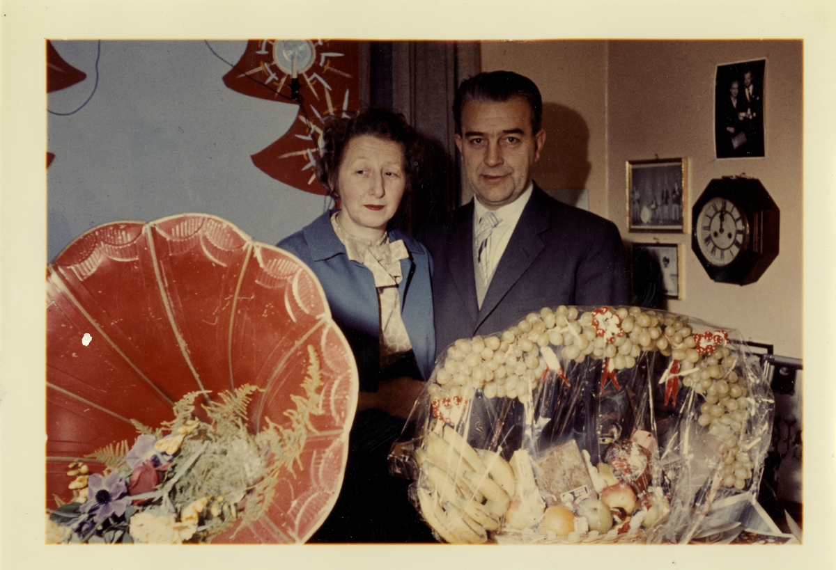 Fotografi av Olaf og Gudrun, ukjent anledning. En stor fruktkurv ses foran i bildet, og en blomsterdekorert grammofontrakt. Sannsynligvis tatt i butikkens andre etasje.