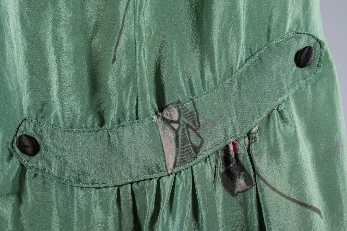 En grønn silkekjole med rosa blomstermønster i Charleston-fasong. Den har lange ermer og V-ringet halsåpning. Ermene er i krepp.chiffon. Mansjettene er smale med trykknapper. Det er svarte stofftrukne pynteknapper i halsåpningen. På sidene er stoffet rynket med påsydd spensel og 2 stofftrukne pynteknapper i sort.