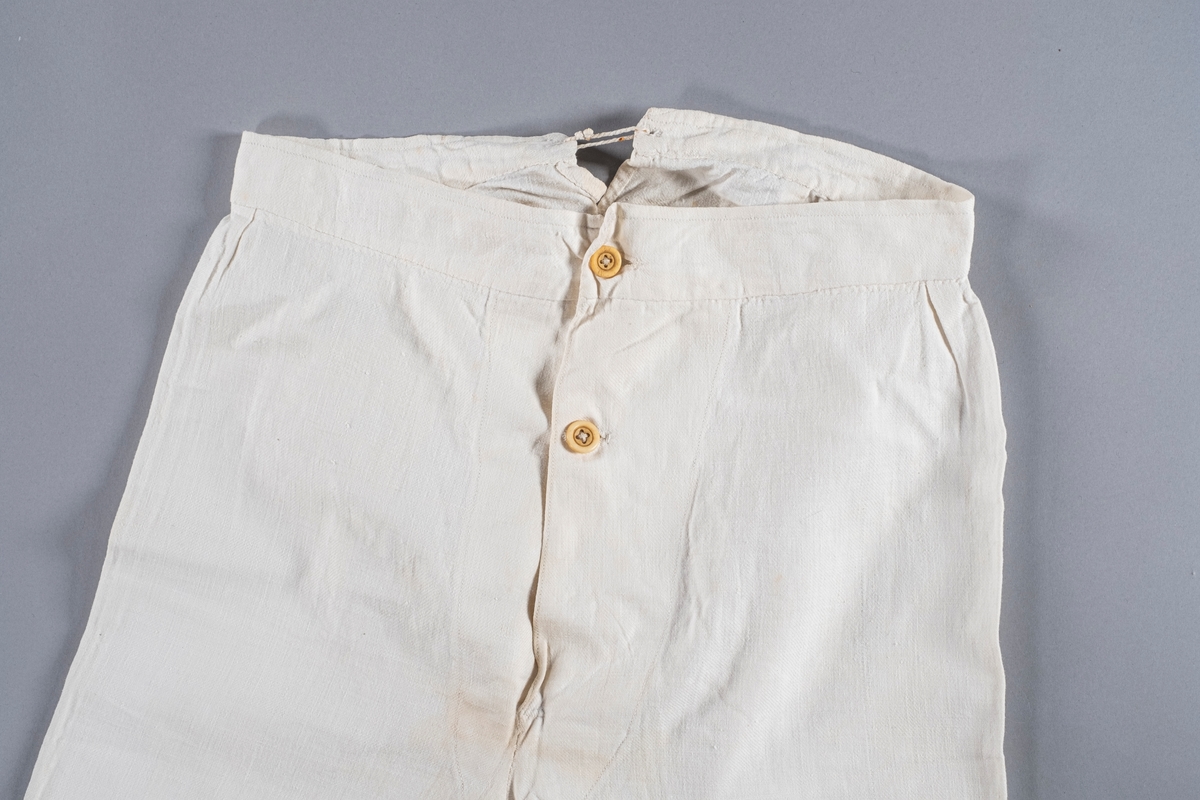 Underbukse i hvit bomull, åpning i front med to knapper, og åpning bak med snøring. Den har folder ved linningen. Nederst i beina er det splitt.