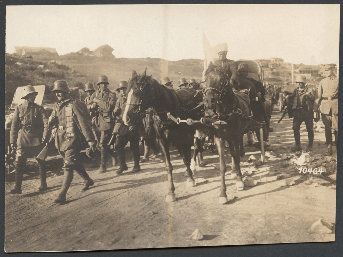 Bilden visar en hästfordon som passerar en kolonn marscherande soldater på en landsväg. På hästvagnen sitter två män kläd i pälsmössa som medför vita flaggan.