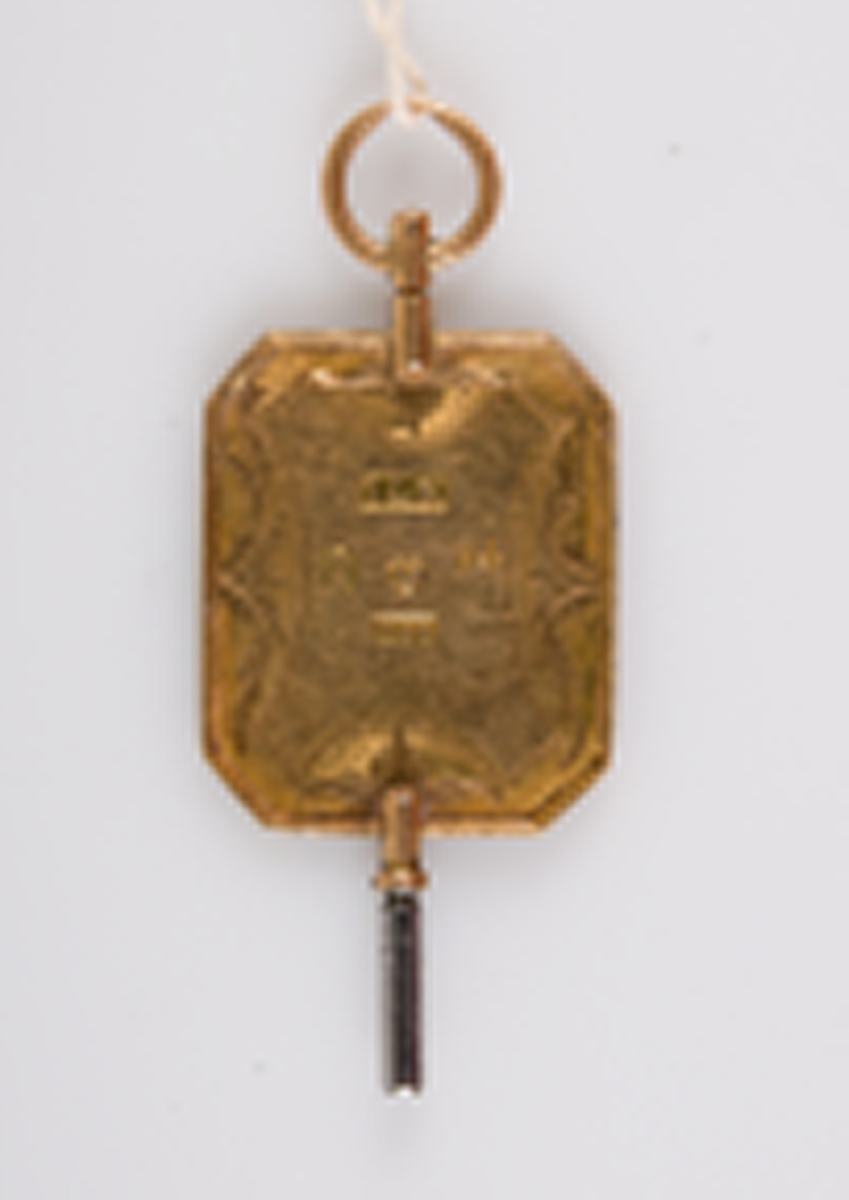 Urnyckel av guld, med åttkantig skiva vars ena sida är försedd med graverad dekor i form av uppslagen notbok, trumpet, kvistar m.m. Stämplar på motsatta sidan: ILK, ankare, kattfot, F 4, 18 K.