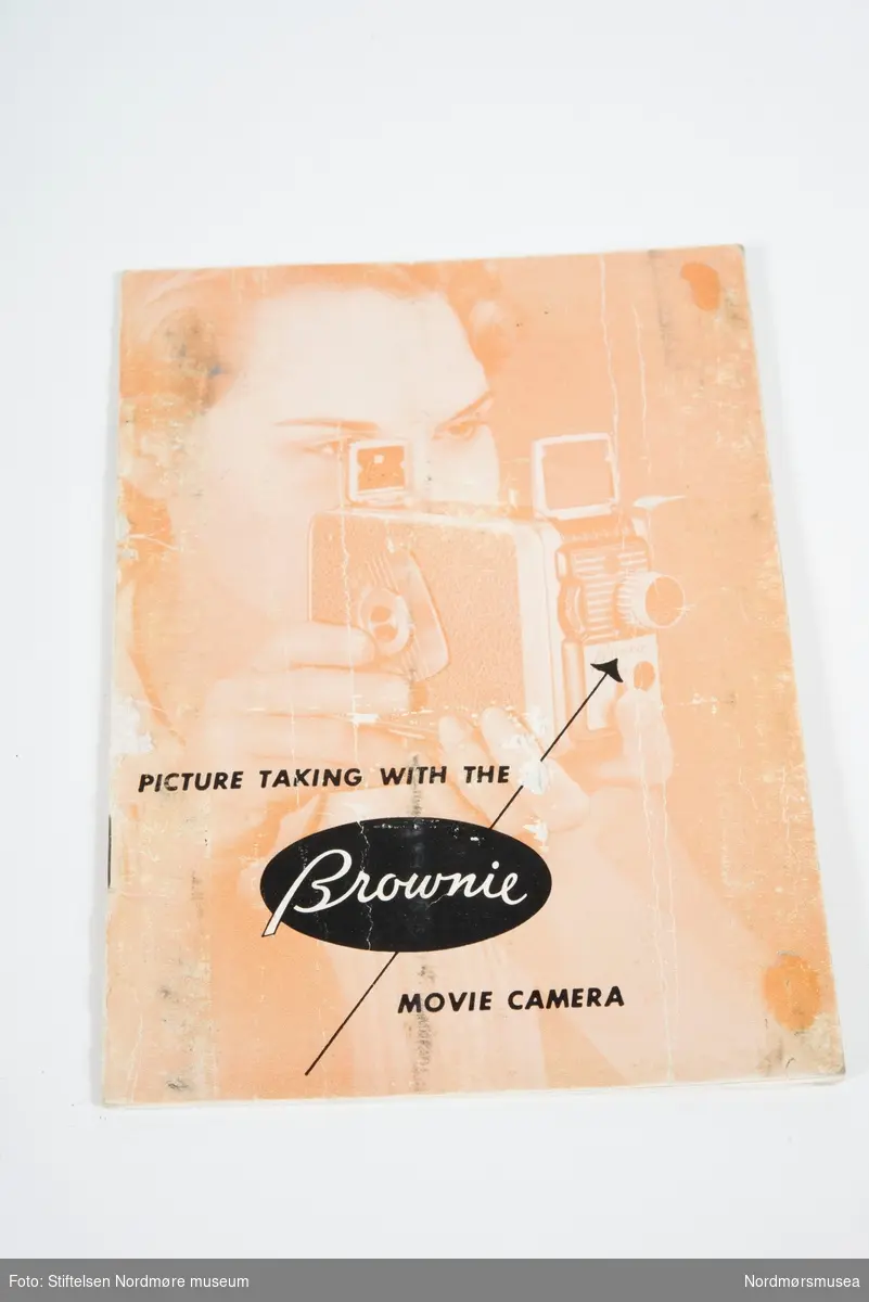 Filmkamera med orginalemballasje. Kameraet er av typen Brownie 8mm og er ment til å brukes med Kodachrome film. Pålimt brunt lær på sidene og den har en profilert plastlist som går rundt hele kamerahuset. Medfølger en liten jukselapp med instrukser på bruk av blenderåpning på høyre side. Manual ligger oppi esken.