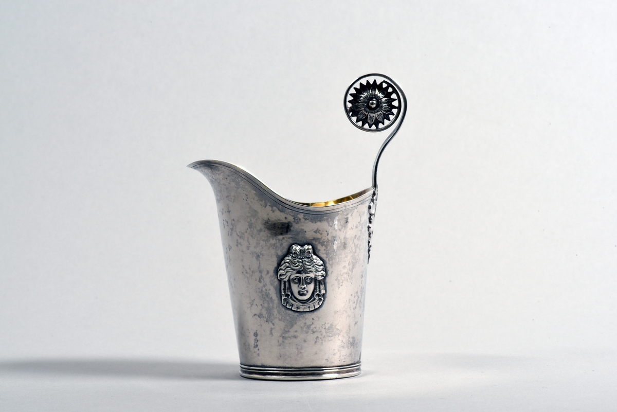 Hjälmformad gräddkanna med handtag av silver. Cuppan är prydd med två ansikten i relief, handtaget är räfflat och utformat som en stiliserad stjälk med blad vid fästet och en blomma högst upp. Insidan är förgylld.
