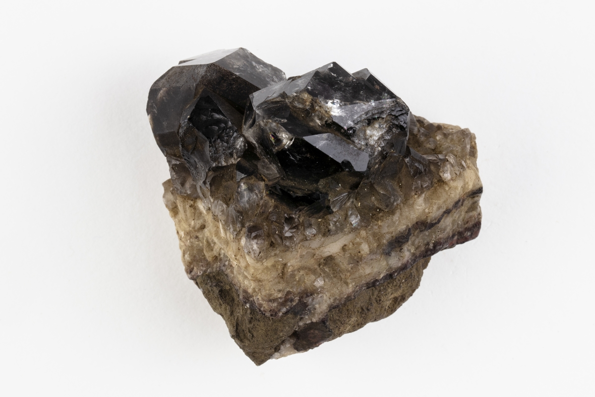Ett mineral som är en kvarts och består av kiseldioxid. Kvartsen har kristallform och färgen är mörk, rökfärgad. Exemplaret kommer från Hässelkulla i Närke, Sverige.