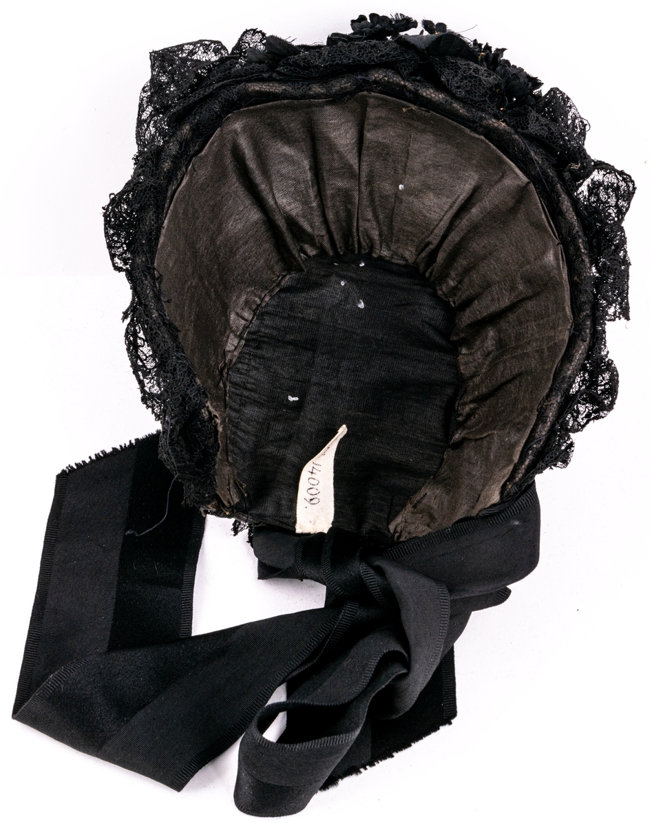 Kapotthatt, svart flätad med tagel och spånband, prydd med spetsar och blombukett, svart med förgyllda ståndare. Svarta knytband av rips.