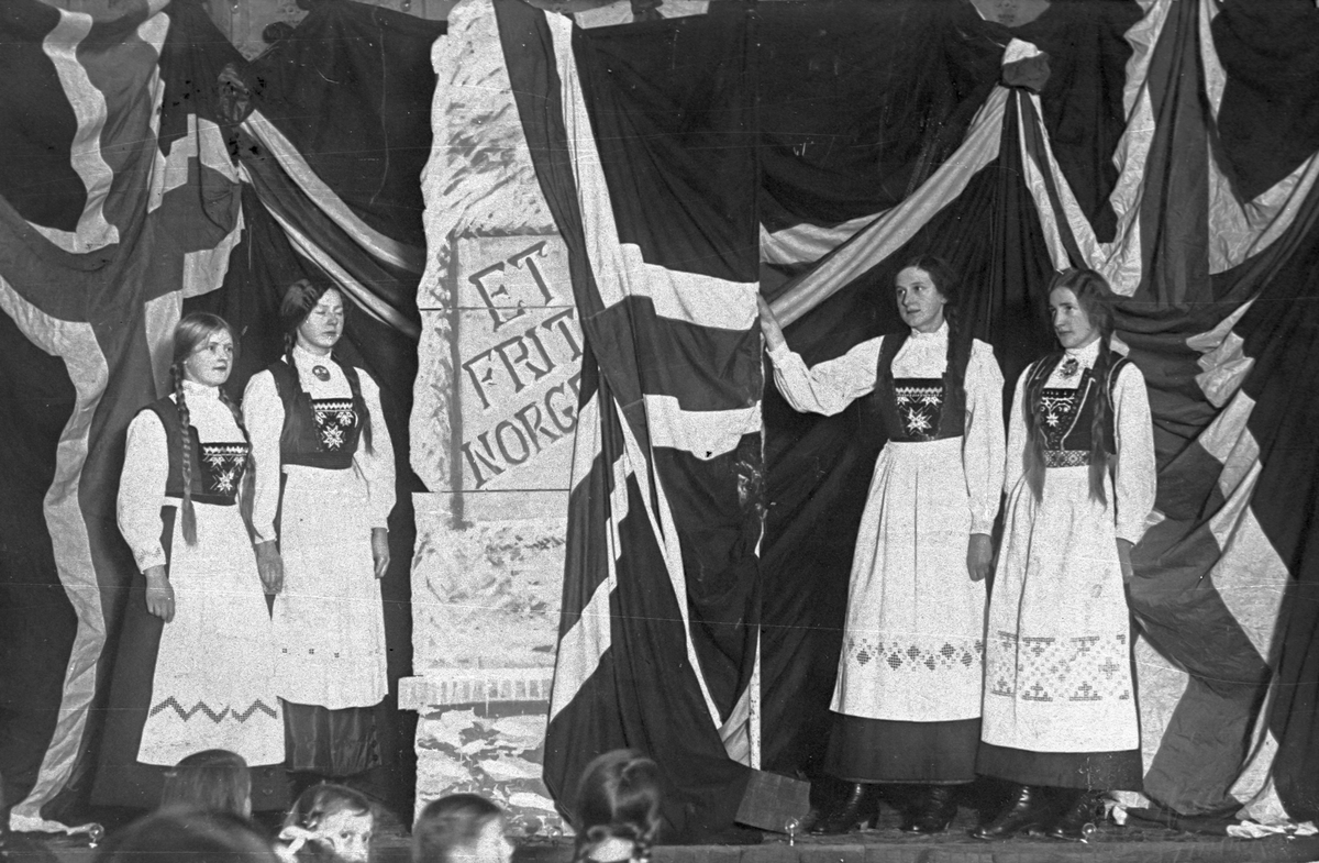 Kvinner i bunad med flagg og faner på ein scene.