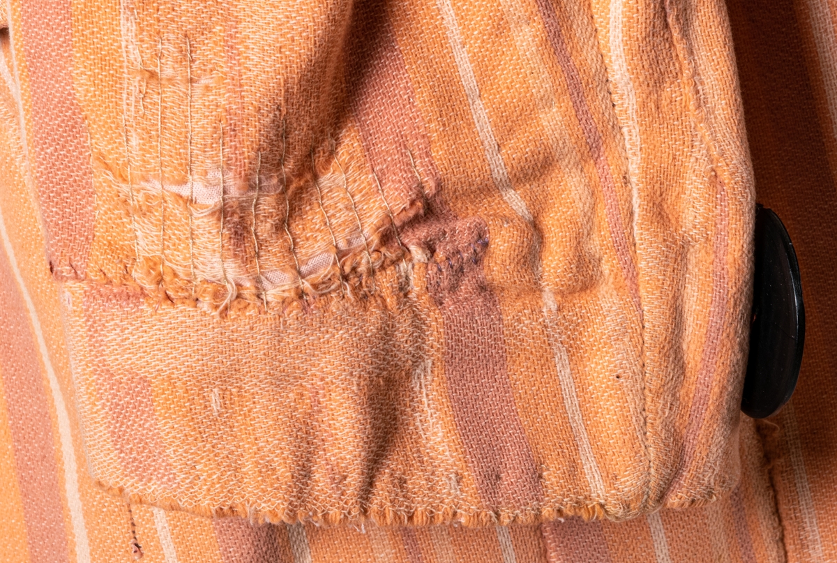 Rock av bomullsflanell mönstrad med vertikala ränder i olika skära toner. Närmast jacketskuren. Krage, slag och tvärskurna sidofickor med ficklock. Falsk ficka på högra skörtets främre del. Knäppes med en stor svarvad knapp. Två dylika i ryggen samt en mindre på var ärn. Enfärgat orange foder, lappat och delvis trasigt.
Mått: ärmlängd 380 mm.
