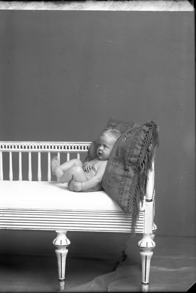 Ett naket spädbarn ligger mot en kudde i en soffa av gustaviansk stil. Det är troligen Åke Rahm.