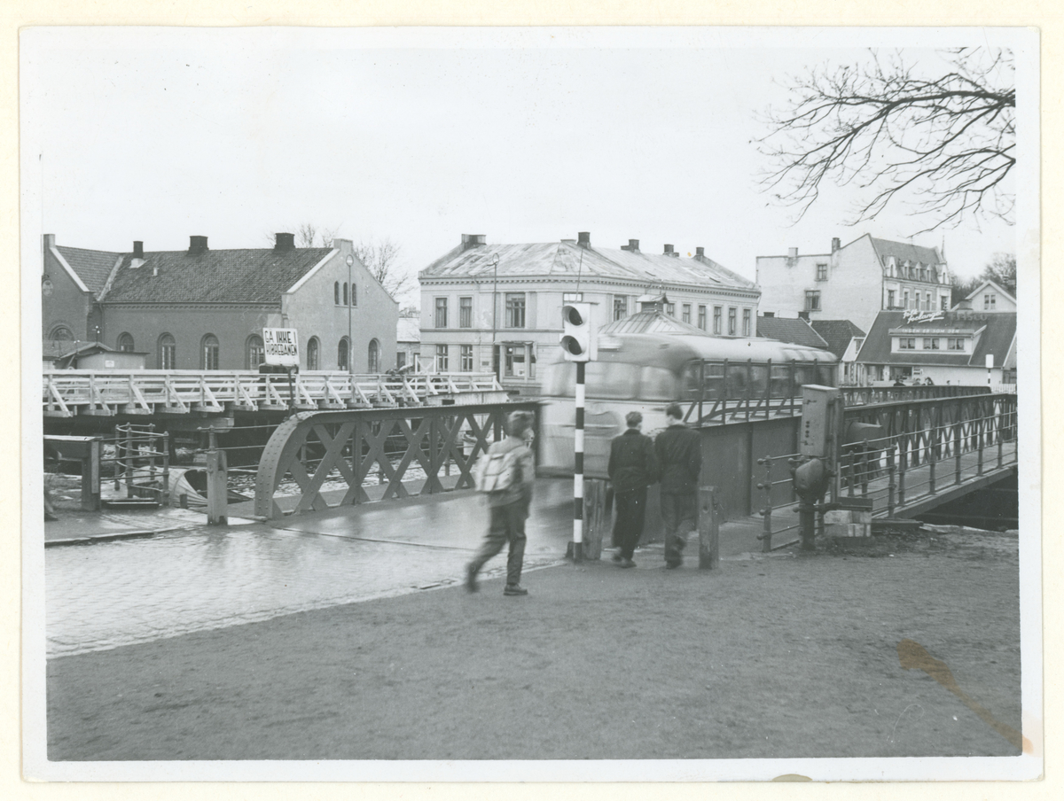 Fotograf har stått ved inngangen til kanalparken på Jeløysiden.
Detaljer: Den midlertidige broen er under oppføring, Broen fra 1888, Tollboden, bygninnger i Jeløygata, Trafikkbrulys, gangbru, folk.
Historikk: Den midlertidige broen sto ferdig i 1955 og var i virke til ny bru var i virke i 1957.