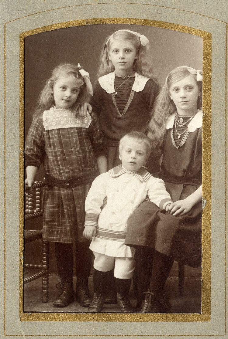 Fyra okända syskon (?), tre flickor och en pojke. Två av flickorna har likadana mörka klänningar, den tredje är klädd i rutigt.
Pojken är klädd i ljus dräkt med mörka garneringar. 
Helfigur. Ateljéfoto.